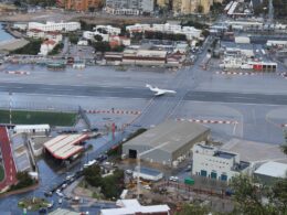 Aerial view of a rainy airport with a white jet on a taxiway and surrounding urban buildings and a stadium track nearby