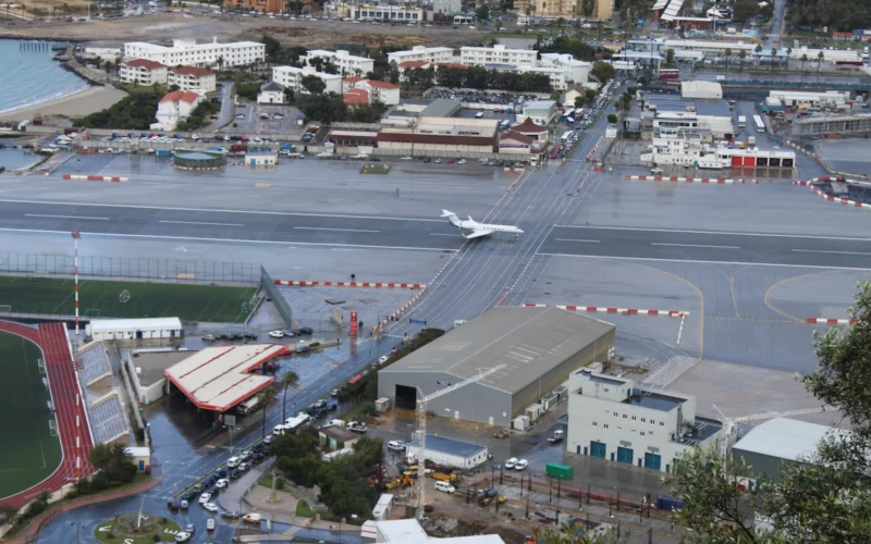 Aerial view of a rainy airport with a white jet on a taxiway and surrounding urban buildings and a stadium track nearby