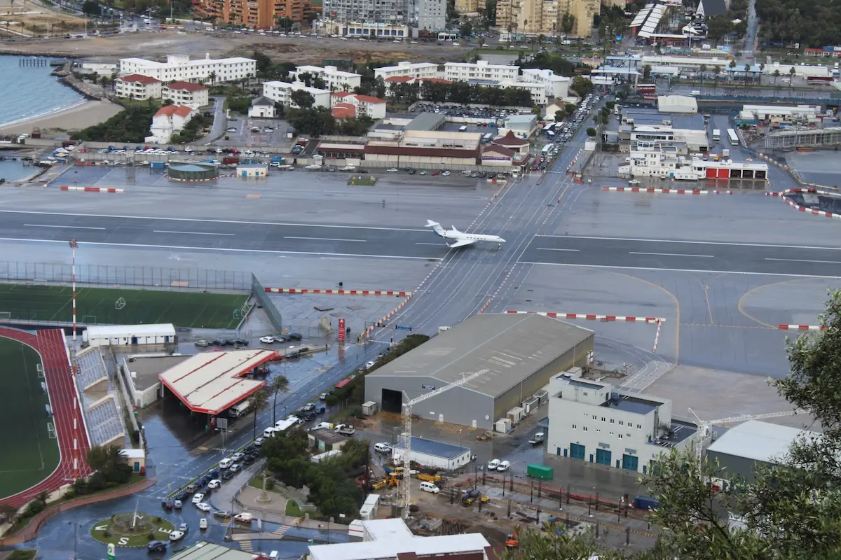 Aerial view of a rainy airport with a white jet on a taxiway and surrounding urban buildings and a stadium track nearby