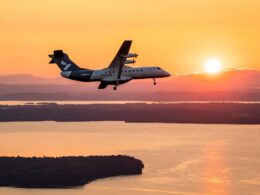 Twin turboprop airplane flying at sunset over a calm body of water with distant land and hills silhouetted beyond the water surface