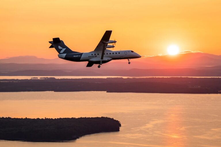 Twin turboprop airplane flying at sunset over a calm body of water with distant land and hills silhouetted beyond the water surface