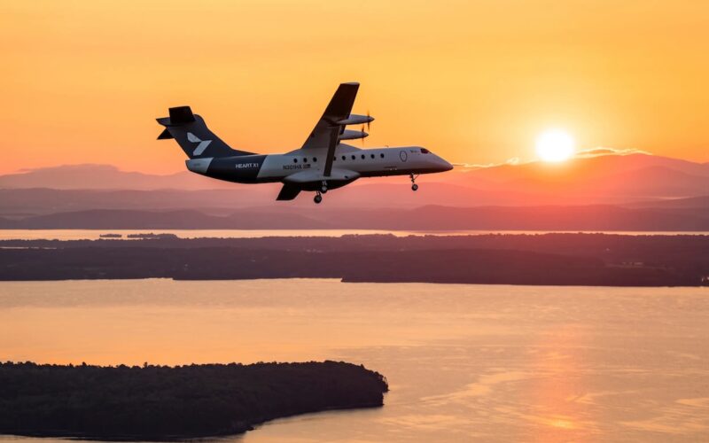 x1 first flight Twin turboprop airplane flying at sunset over a calm body of water with distant land and hills silhouetted beyond the water surface