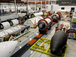Inside a large aerospace factory with rocket modules on conveyors and workstations including a black nose cone on a yellow cradle in the foreground and red cylindrical stages in the background