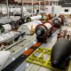 Inside a large aerospace factory with rocket modules on conveyors and workstations including a black nose cone on a yellow cradle in the foreground and red cylindrical stages in the background