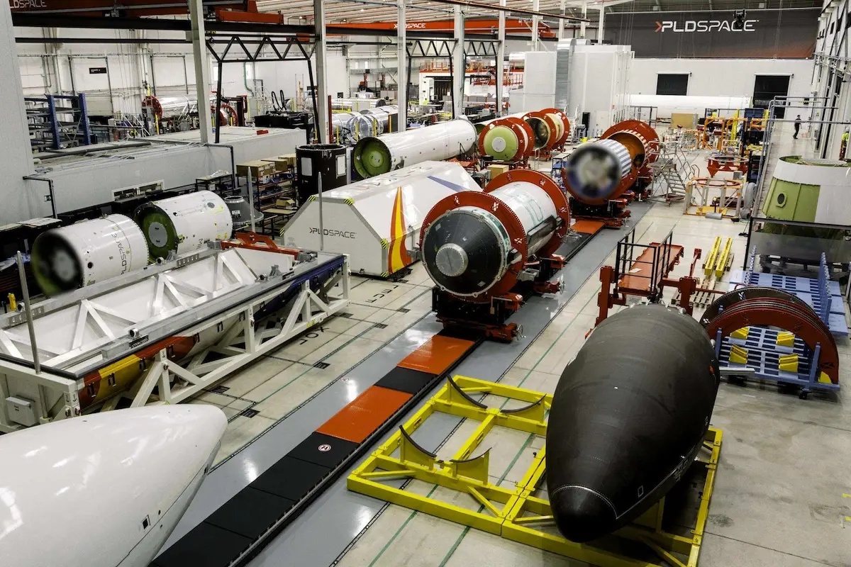 Inside a large aerospace factory with rocket modules on conveyors and workstations including a black nose cone on a yellow cradle in the foreground and red cylindrical stages in the background