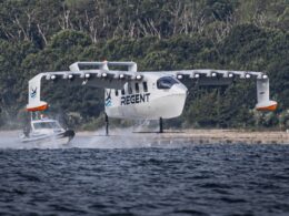 Regent seaplane landing on a lake with a support boat creating spray white aircraft with two winged pontoons and REGENT on the fuselage near a tree lined shore