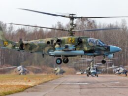 A Russian Ka 52 Alligator attack helicopter hovers above an airfield with other Ka 52 and Mi 28 helicopters in the background