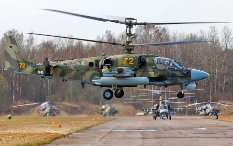 A Russian Ka 52 Alligator attack helicopter hovers above an airfield with other Ka 52 and Mi 28 helicopters in the background