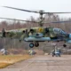 A Russian Ka 52 Alligator attack helicopter hovers above an airfield with other Ka 52 and Mi 28 helicopters in the background