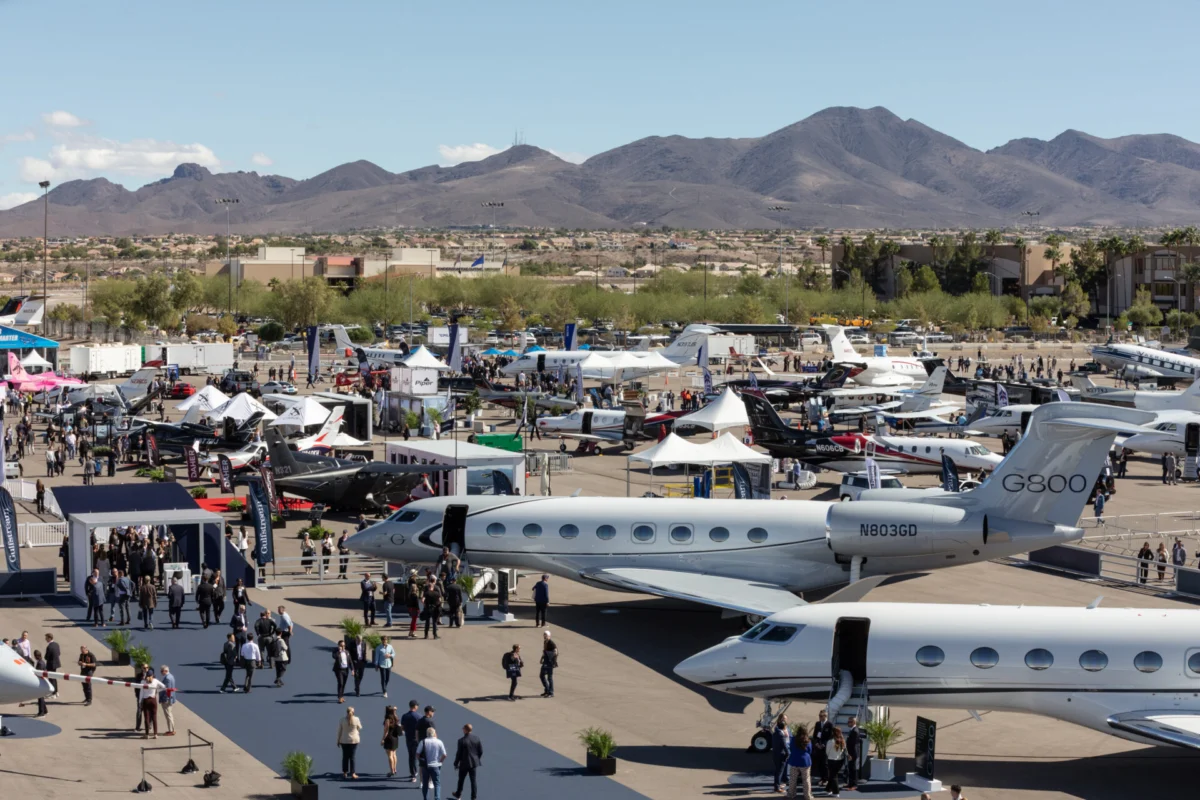 Aircraft exhibited at NBAA BACE Air show at an airport with numerous private jets tents and attendees set against desert mountains in the distance