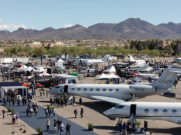 Air show at an airport with numerous private jets tents and attendees set against desert mountains in the distance