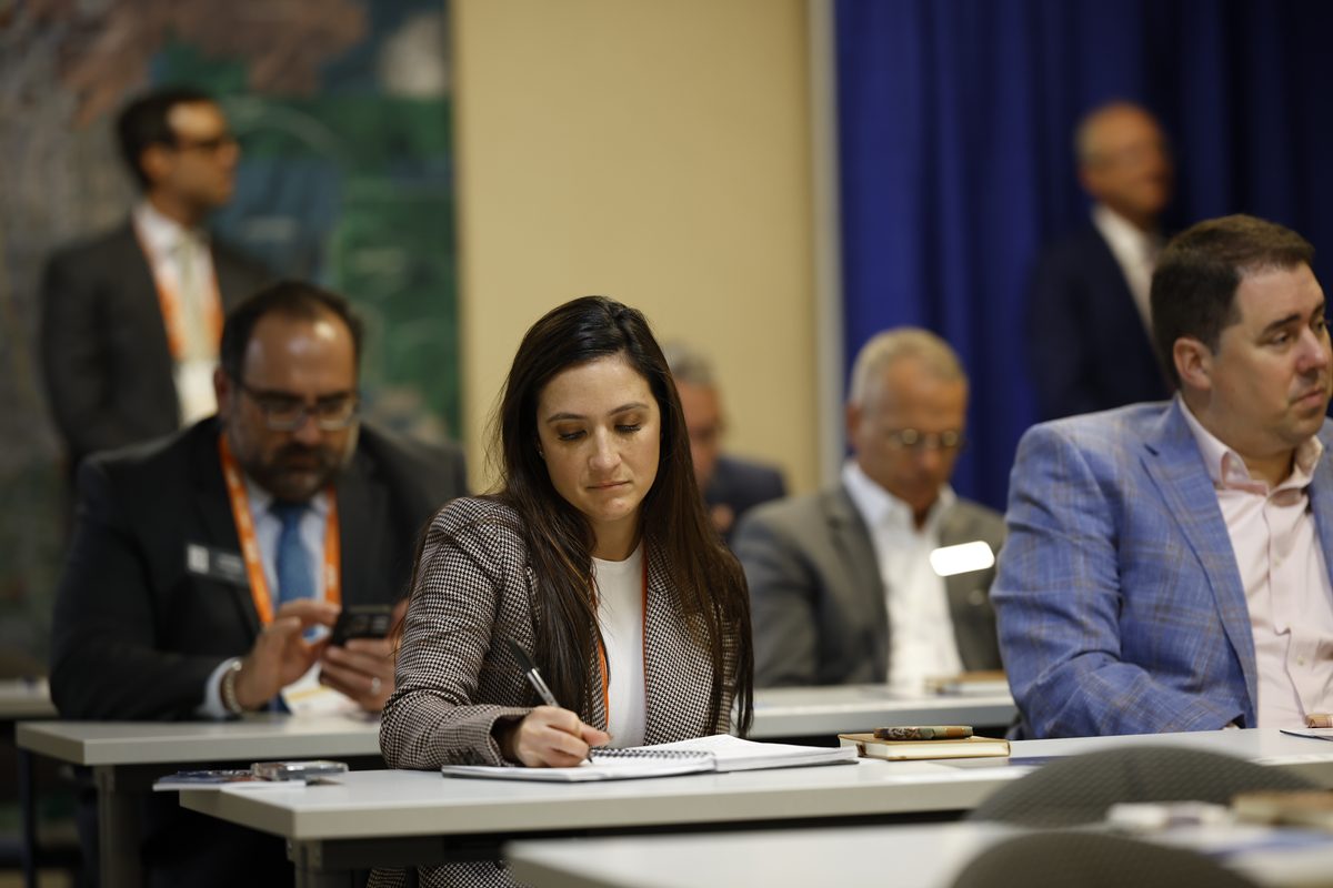 Woman in a blazer writes in a notebook at a conference table colleagues blurred in the background