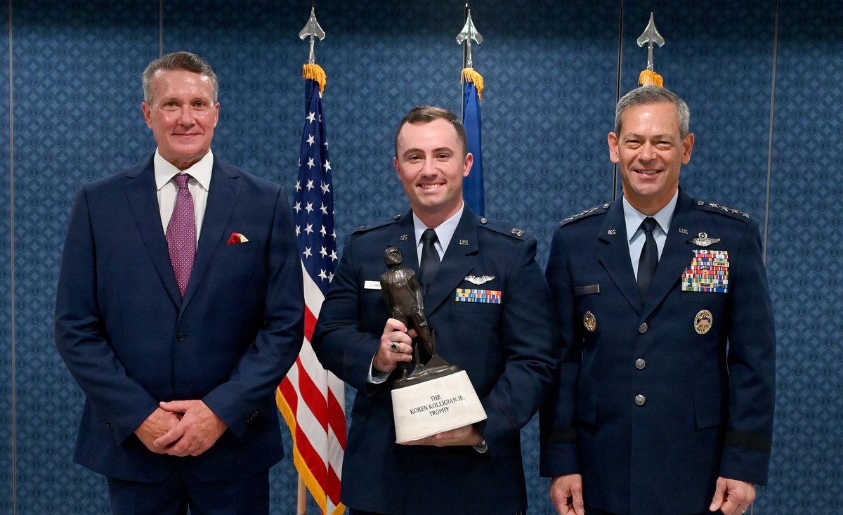 90th Fighter Squadron Captain receives 2025 Koren Kolligian Jr Trophy Three men pose for a formal award photo a civilian on the left in a navy suit two uniformed officers on the right the middle one holding a trophy with flags in the background