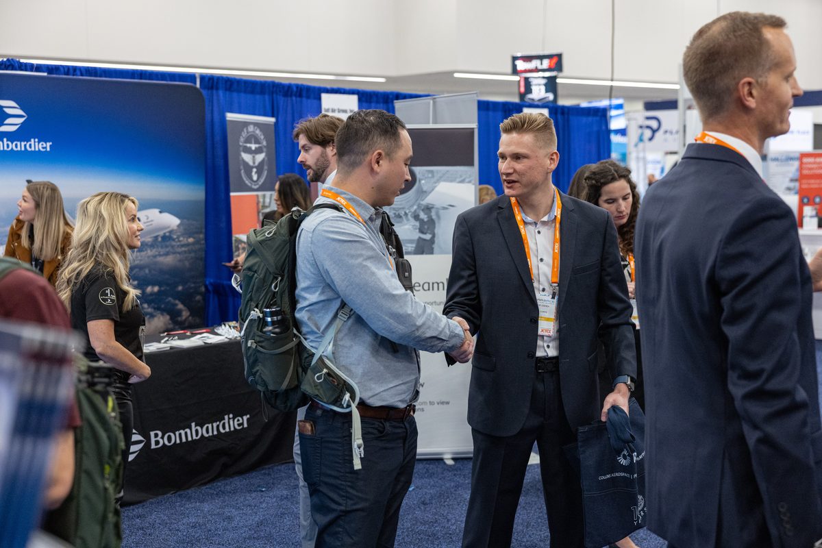 Two men shake hands at a crowded trade show booth with Bombardier branding in the background