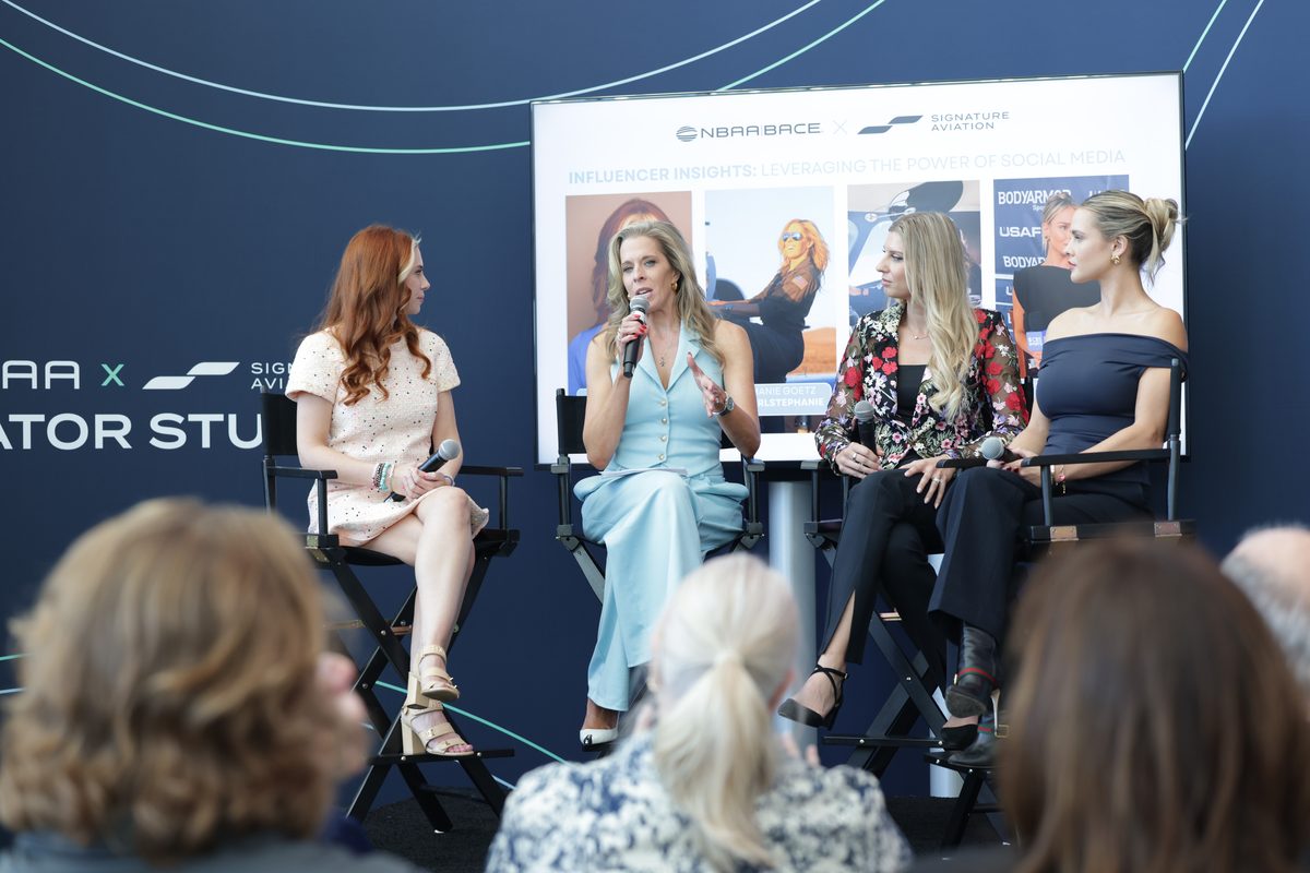 Panel of four women seated on stage in a discussion audience visible in foreground dark blue backdrop with logos and screen behind them