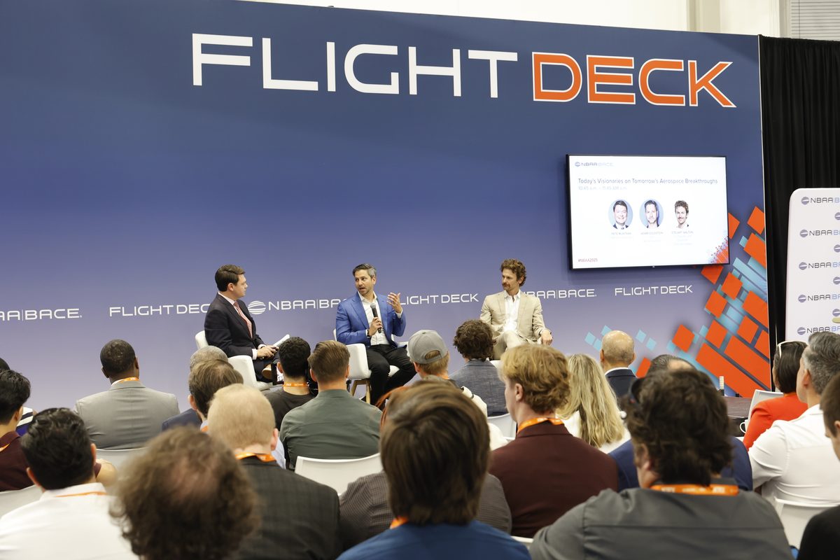 Panel discussion on a blue stage with a large FLIGHTDECK sign three men in suits seated and speaking to an audience