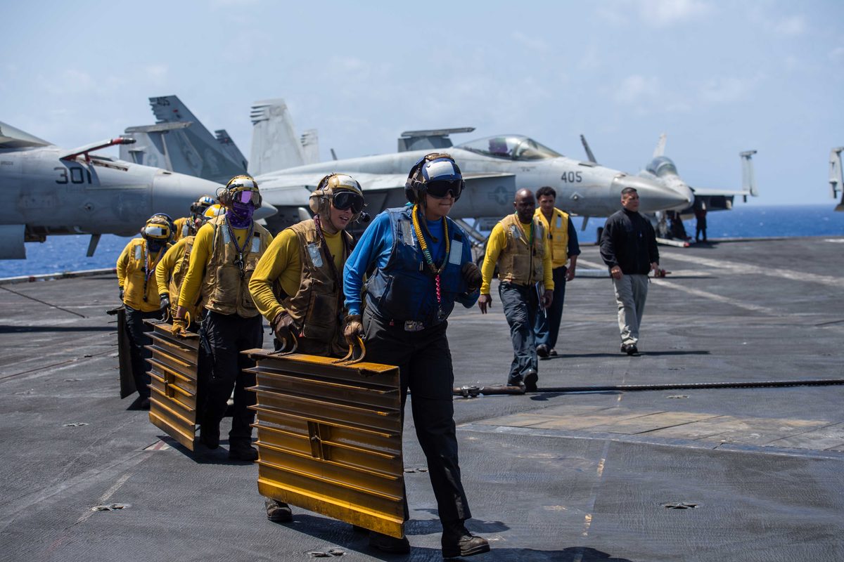 USS Dwight D Eisenhower CVN 69 Conducts Flight Deck Drills in the Mediterranean Sea Sailors in yellow vests and helmets carry folded metal panels across an aircraft carrier deck with fighter jets parked in the background