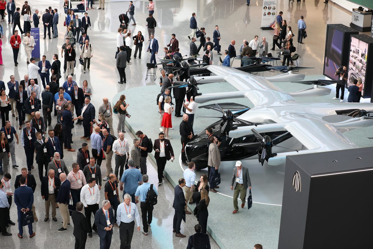 Crowded conference hall with attendees wearing badges networking near a row of silver aircraft on display at an aerospace expo
