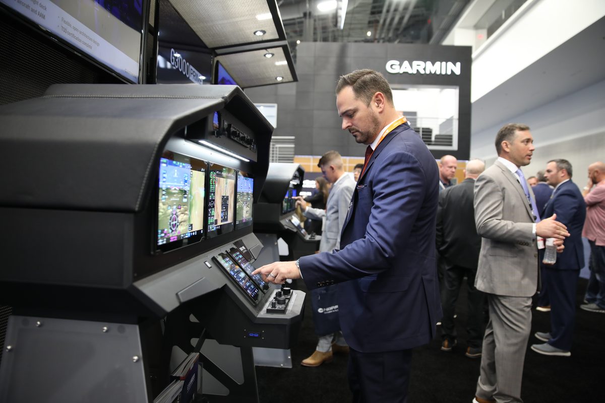 Man in a navy suit uses a large aircraft cockpit simulator at a trade show with screens and other attendees in the background Garmin booth visible