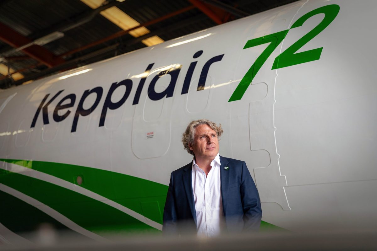 Portrait David Joubert Pdt Kepplair Evolution©KepplairEvolution Businessman in a navy suit stands beside a large white airplane with bold dark lettering and green accents inside a hangar