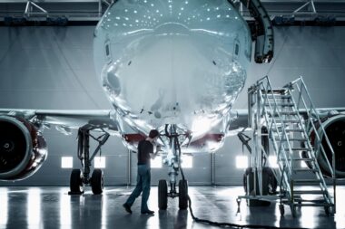 Technician inspecting the nose of a large jet in a hangar with mobile stairs nearby during maintenance