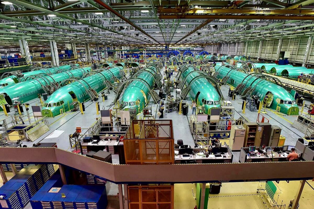 Wide view of an airplane assembly plant with multiple green fuselage sections on the production line workers and equipment visible throughout