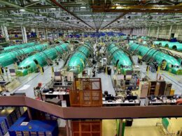Wide view of an airplane assembly plant with multiple green fuselage sections on the production line workers and equipment visible throughout