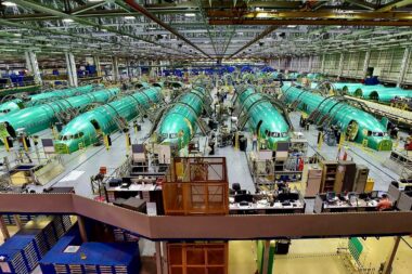 Wide view of an airplane assembly plant with multiple green fuselage sections on the production line workers and equipment visible throughout