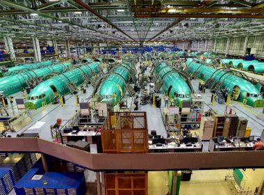 Wide view of an airplane assembly plant with multiple green fuselage sections on the production line, workers and equipment visible throughout.