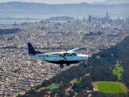 Small white and blue aircraft flying low over a densely built city with hills and a bay in the background