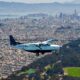 Small white and blue aircraft flying low over a densely built city with hills and a bay in the background