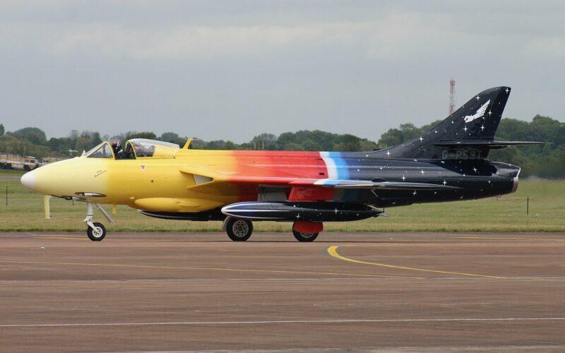 Recently Saved 1 of 1 15 Side view of a colorful aerobatic jet on a taxiway yellow nose rainbow gradient along the fuselage and a starry dark tail with a white emblem