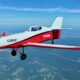Small red and white single engine aircraft in flight over countryside propeller blurred from speed cockpit visible with pilot inside