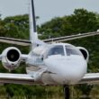 Front view of a white private jet with side mounted engines landing gear down taxiing on a runway with green trees in the background