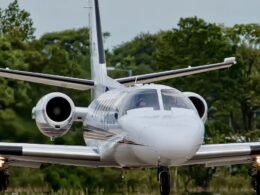 Front view of a white private jet with side mounted engines landing gear down taxiing on a runway with green trees in the background