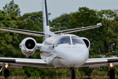 Front view of a white private jet with side mounted engines landing gear down taxiing on a runway with green trees in the background