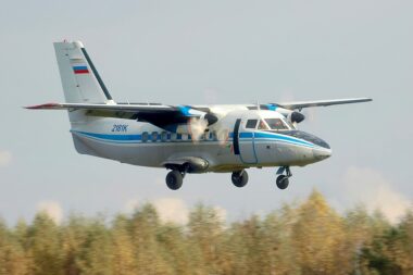 Twin turboprop airplane with landing gear down approaching runway over a treed landscape white with blue stripes and Russian flag on tail