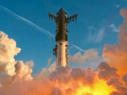 Rocket lifting off through a plume of orange and yellow flames and smoke rising into a blue sky with clouds around it