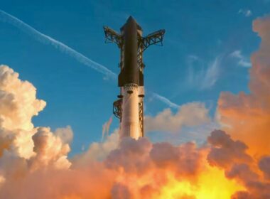 Rocket lifting off through a plume of orange and yellow flames and smoke, rising into a blue sky with clouds around it.