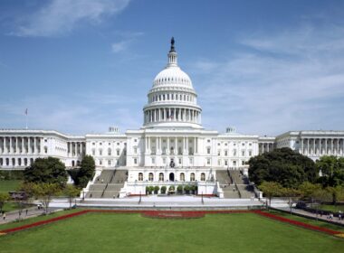 Front view of the U.S. Capitol with its large central dome, wings on both sides, and a manicured lawn in the foreground.
