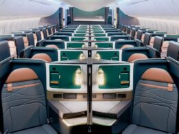 Wide view of a modern airplane cabin with private business class suites teal storage panels and peach seats along a long aisle
