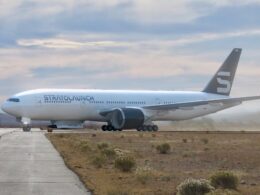 Large white commercial airliner on a taxiway in a desert airfield with a dark tail fin bearing a light logo and distant mountains in the background