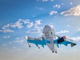 Blue and white experimental aircraft in flight against a clear blue sky with scattered clouds landing gear visible