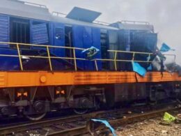 Blue locomotive on an orange railcar appears damaged and derailed with scattered debris along the tracks in a cloudy scene
