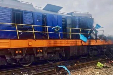 Blue locomotive on an orange railcar appears damaged and derailed with scattered debris along the tracks in a cloudy scene