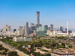 City skyline with a tall glass skyscraper at center against a clear blue sky lower buildings and greenery in the foreground with mountains in the distance