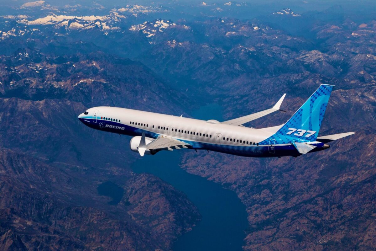 Boeing 737 MAX in blue and white livery flying over a rugged mountain range beneath a clear sky