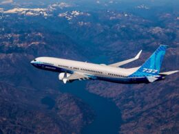 Boeing 737 MAX in blue and white livery flying over a rugged mountain range beneath a clear sky