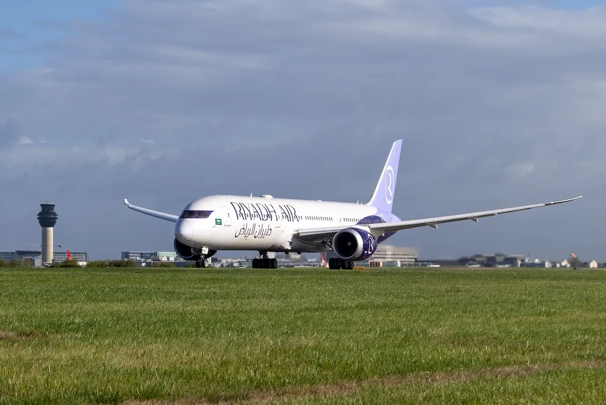 Riyadh aircraft at MAN Riyadh Air widebody airplane taxiing on a runway with a purple tailed livery and a control tower in the distance