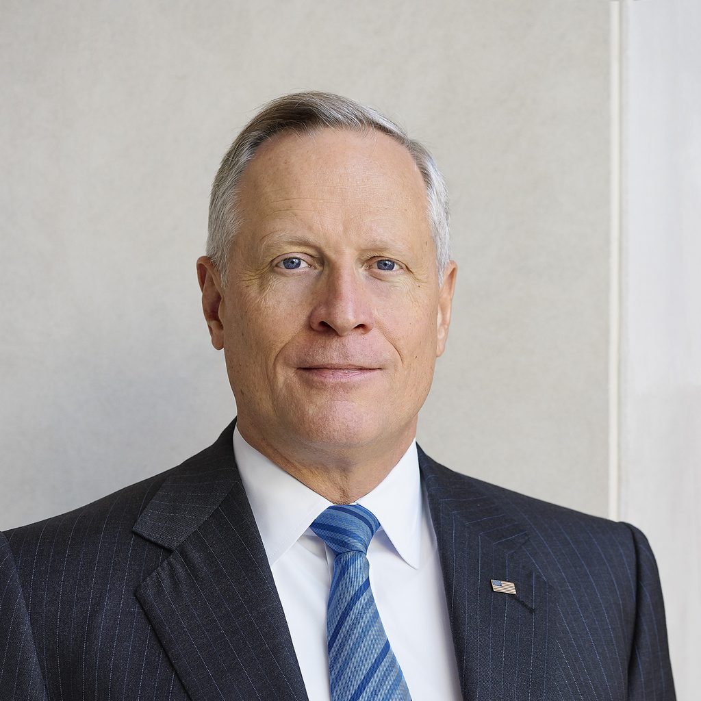 Ross Perot Jr Headshot Portrait of a middle aged man in a dark pinstripe suit white shirt and blue striped tie wearing an American flag lapel pin